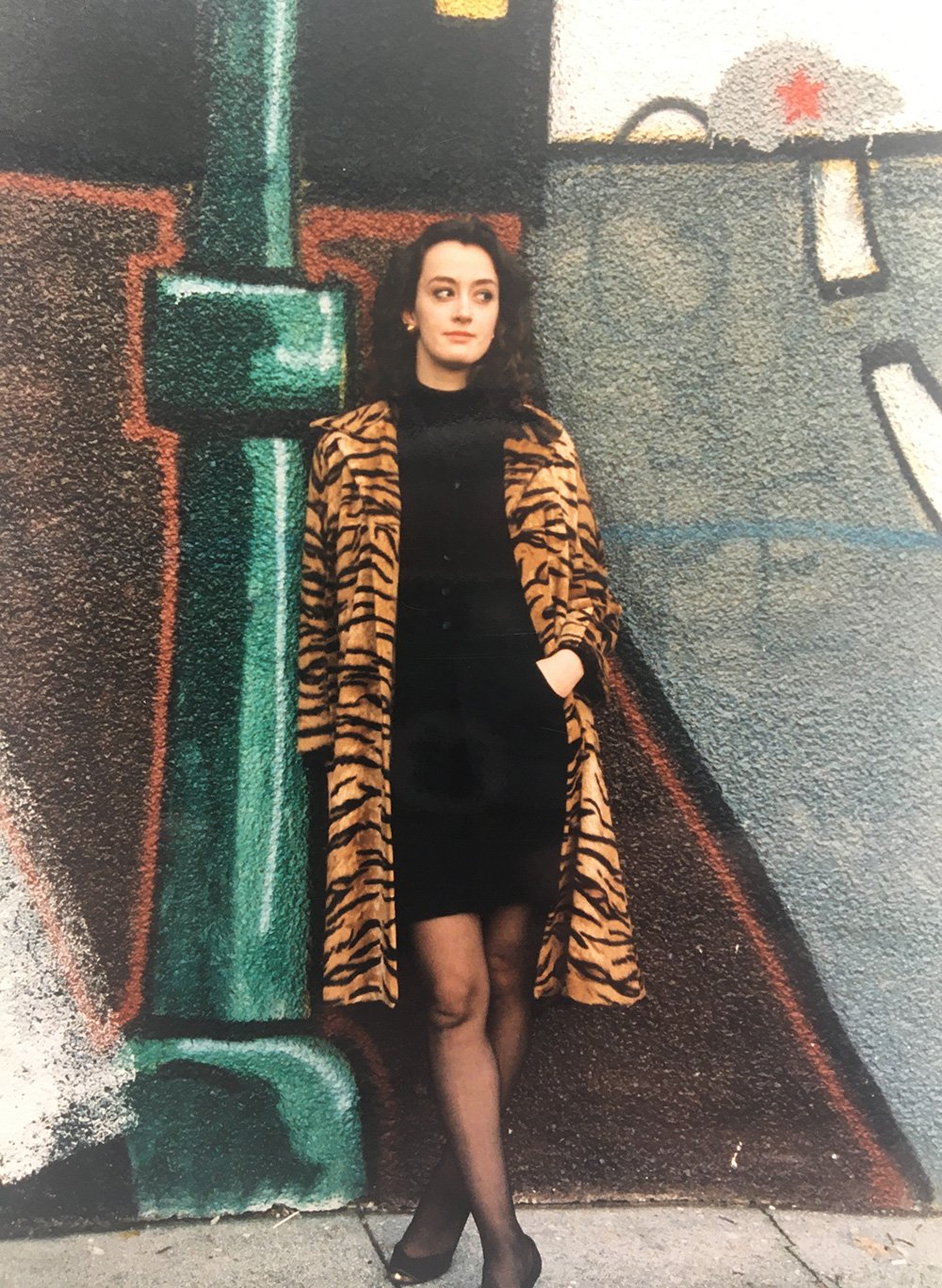 Dorothy Woodend stands against a graffiti wall in a short black dress, long tiger-print jacket, black tights and black shoes. She has long dark hair and is looking to the right.
