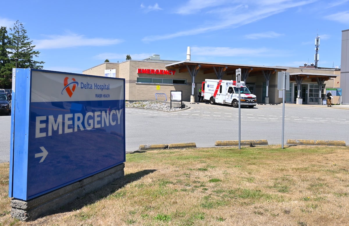 A sign in the foreground reads ‘Delta Hospital. Fraser Health. Emergency.’ In the background, an ambulance is parked in front of a one-story tan brick building with a red ‘Emergency’ sign. 