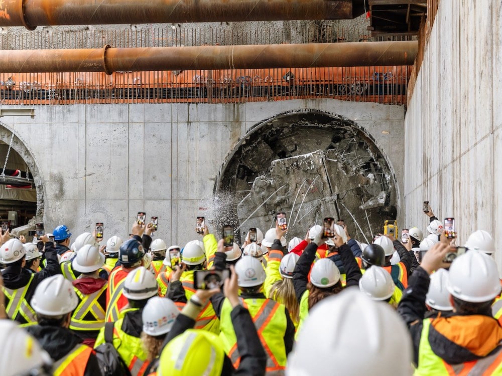 A crowd of people in white hard hats and fluorescent yellow construction vests stand before a grey cement wall into which a tunnel boring machine is breaking through a circular hole. Many members of the crowd are holding up smartphones to take pictures of the hole.