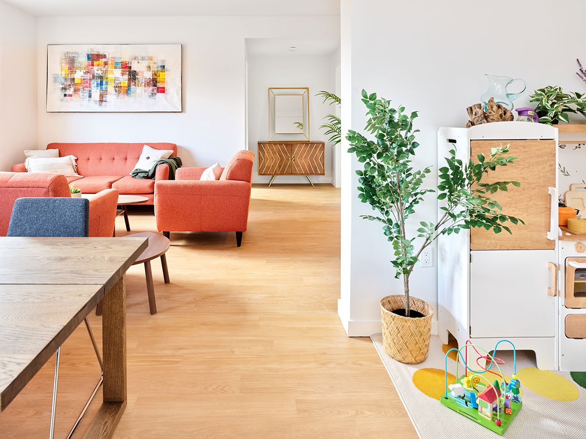 On the right, a wooden child’s kitchen playset and a plant. To the left, farther into the background, comfortable furniture.
