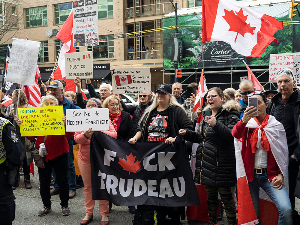 A group of people stand on a Vancouver street, with Canadian flags and signs that say 'Fuck Trudeau' and 'Trudeau must go to prison now.'
