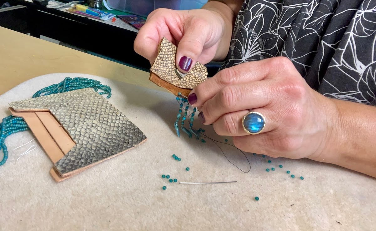 Two hands sew turquoise beads onto a small pouch made of light brown fish leather. An unfinished fish leather wallet lies close by.