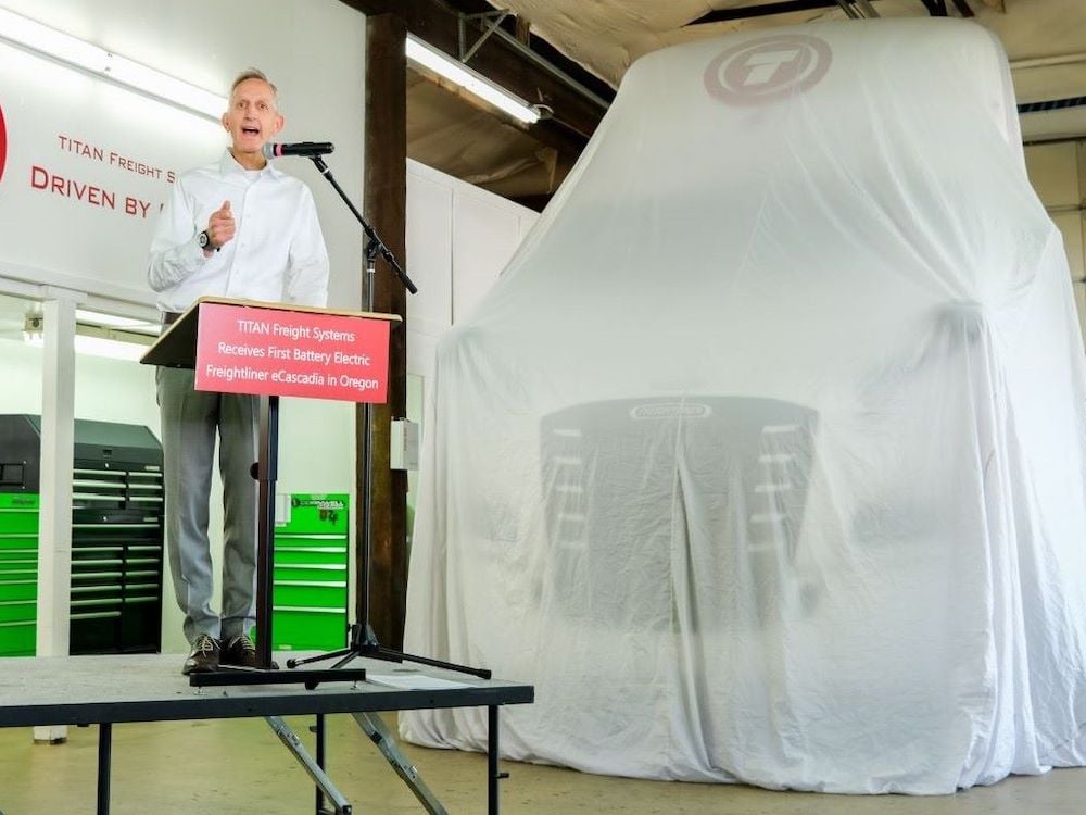 A light-skinned man wearing a white dress shirt and grey slacks stands at a podium with a sign reading 'TITAN Freight Systems Receives First Battery Electric Freightliner eCascadia in Oregon.' Next to him a truck can be made out under a white drop sheet.