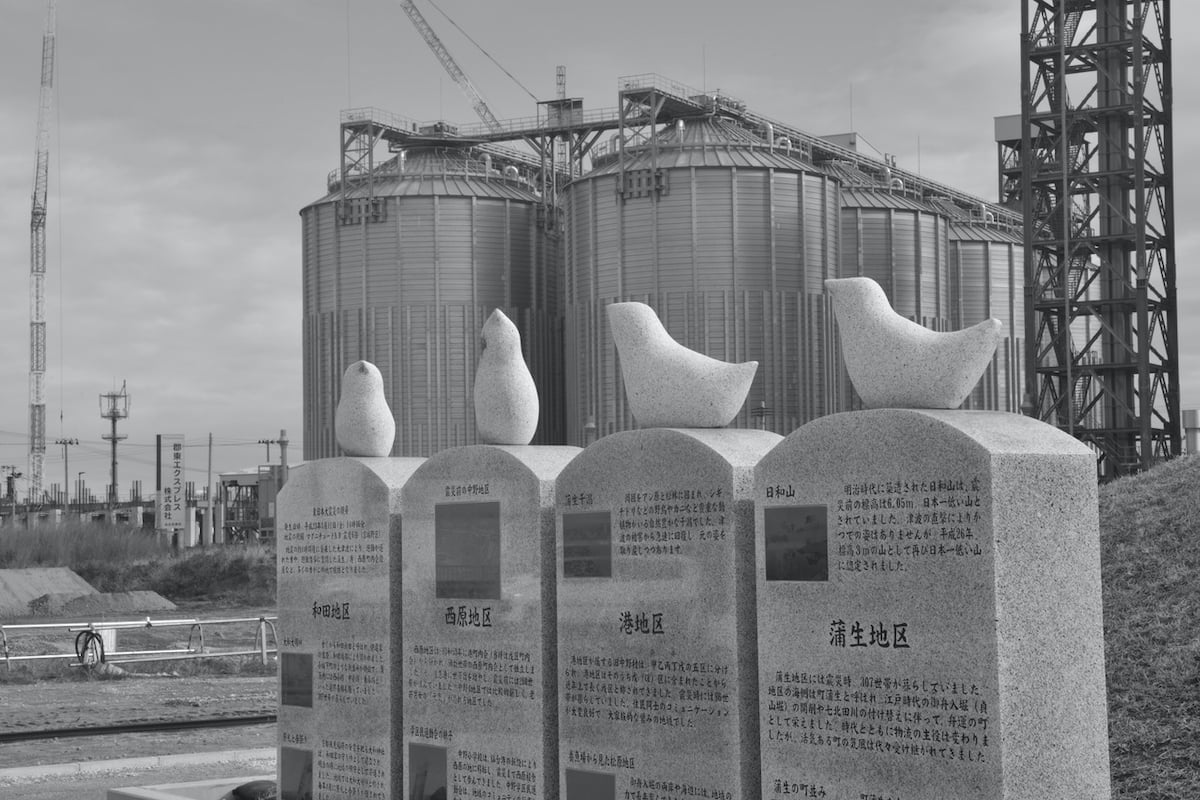 A black and white photo shows headstones with carving; on top of each is a stylized carved bird. In the background are two giant round steel-grey structures.