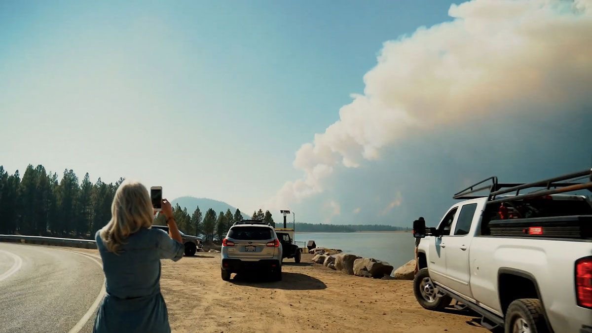A woman with blond hair, a blue T-shirt and light skin holds an iPhone up towards the horizon, where a white plume of wildfire smoke stretches across the blue sky. In front of her are parked sports utility vehicles along a dirt road by the water.