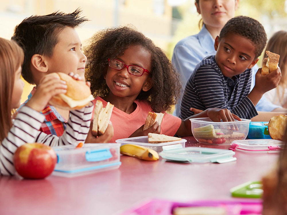 A group of five racially diverse elementary school-aged children sits together at a red table eating lunch.