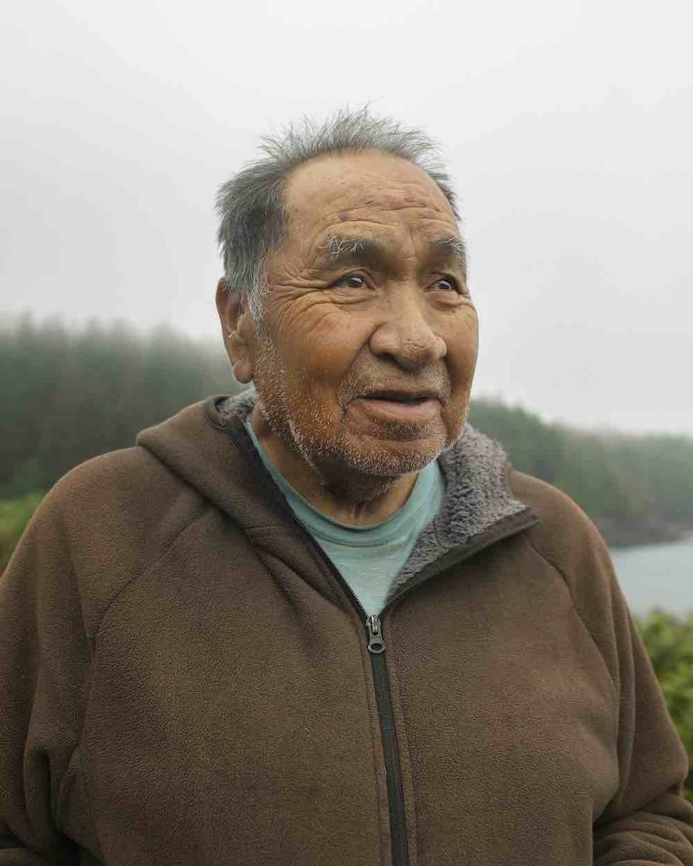 Ray Williams is an elderly Indigenous man with medium skin tone, short dark hair and a brown zip-up hoodie with fleece lining over a turquoise T-shirt. In the soft-focus background is a grey sky and a stand of coniferous trees.