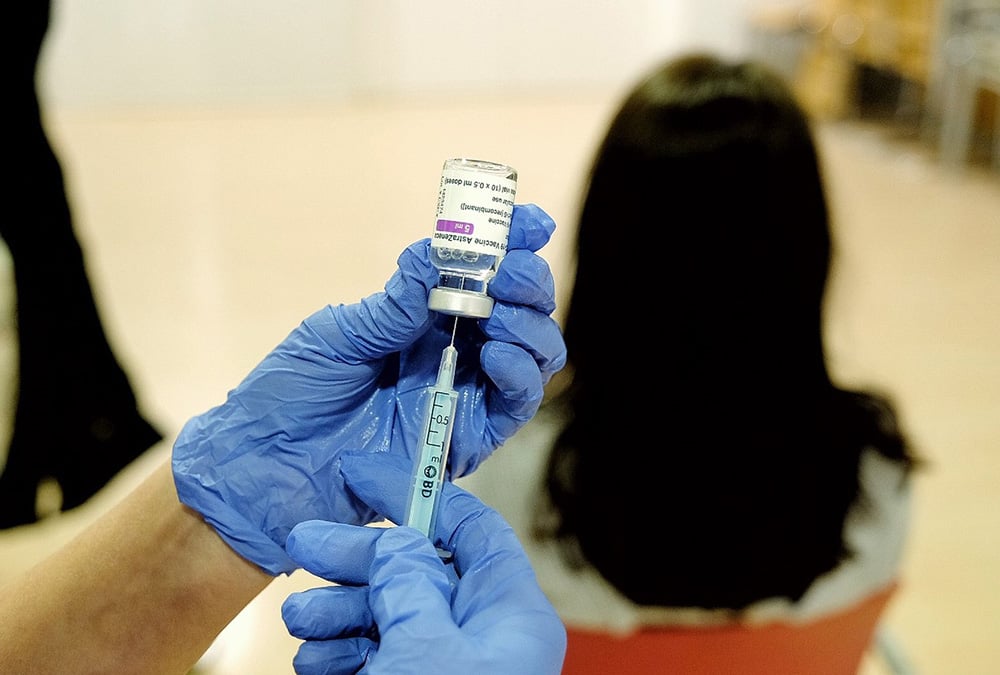 A person wearing blue latex gloves draws a needle full of COVID vaccine from a vial. Another person, blurry, in the background, is sitting, waiting to get their COVID booster.