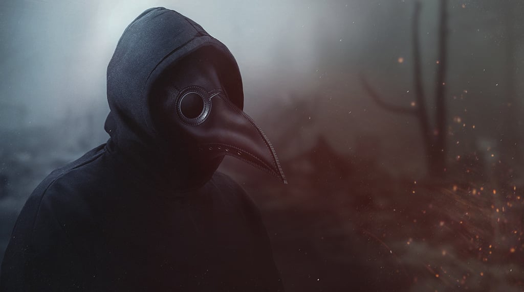 A person wearing a crow mask, the traditional image for medieval plague doctors, and a black hoodie in front of smoke and sparks from a fire.