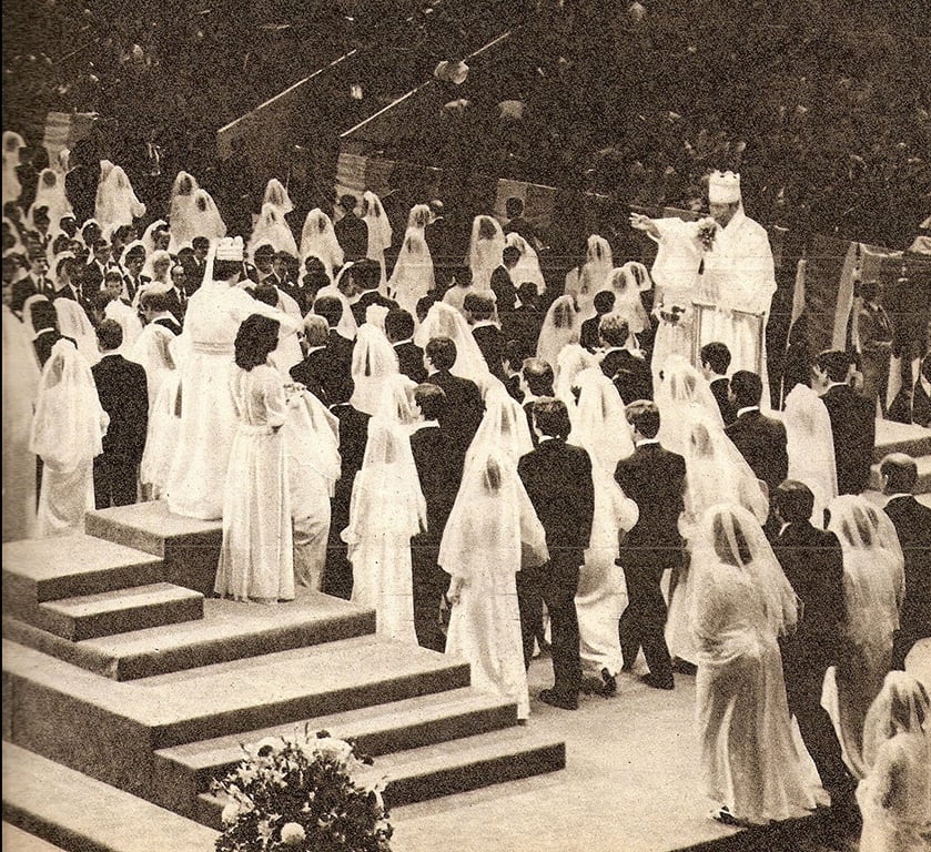 A line of men in dark suits with women in white dresses parade in from of a man wearing robes and crown.