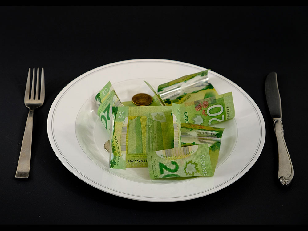 A handful of Canadian $20 bills are crumpled over a white dinner plate. A fork sits on the left and a knife on the right amidst a black background.