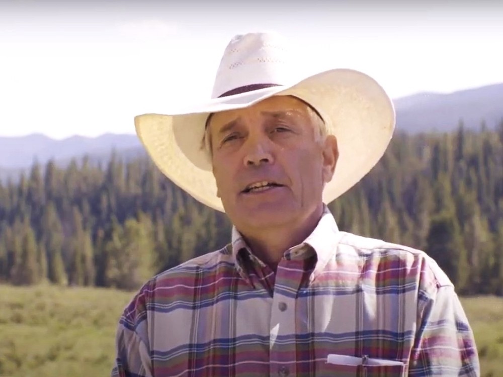Close-up of a man in a cowboy hat and plaid shirt, with a grassy field, trees and hills behind him.