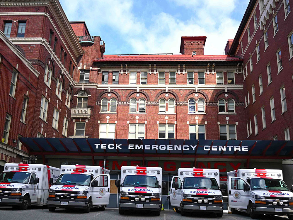 A busy ambulance bay outside the emergency room at St. Paul’s Hospital in Vancouver. Five ambulances are parked in a row in front of a red brick building. 