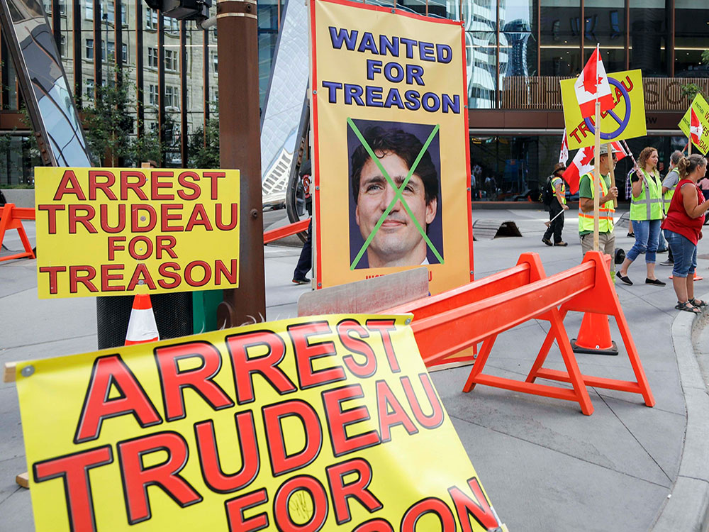 A few protesters, mostly dress in safety vests, stand outside a building. Yellow signs call on Prime Minister Justin Trudeau t be arrested for treason.