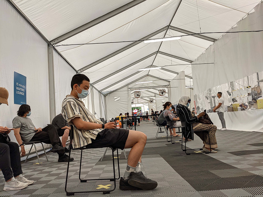 Several people sit on black chairs under a white medical tent with grey flooring. They are all wearing blue medical masks, many of them checking their phones.
