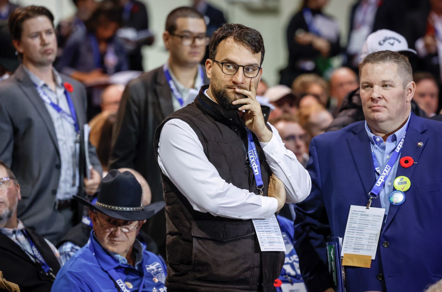A white man with short brown hair and beard, wearing a black vest over a white shirt, holds his hand to his jaw and glances sideways toward the left of the frame. He is surrounded by people wearing lanyards that say 'UCP AGM.'