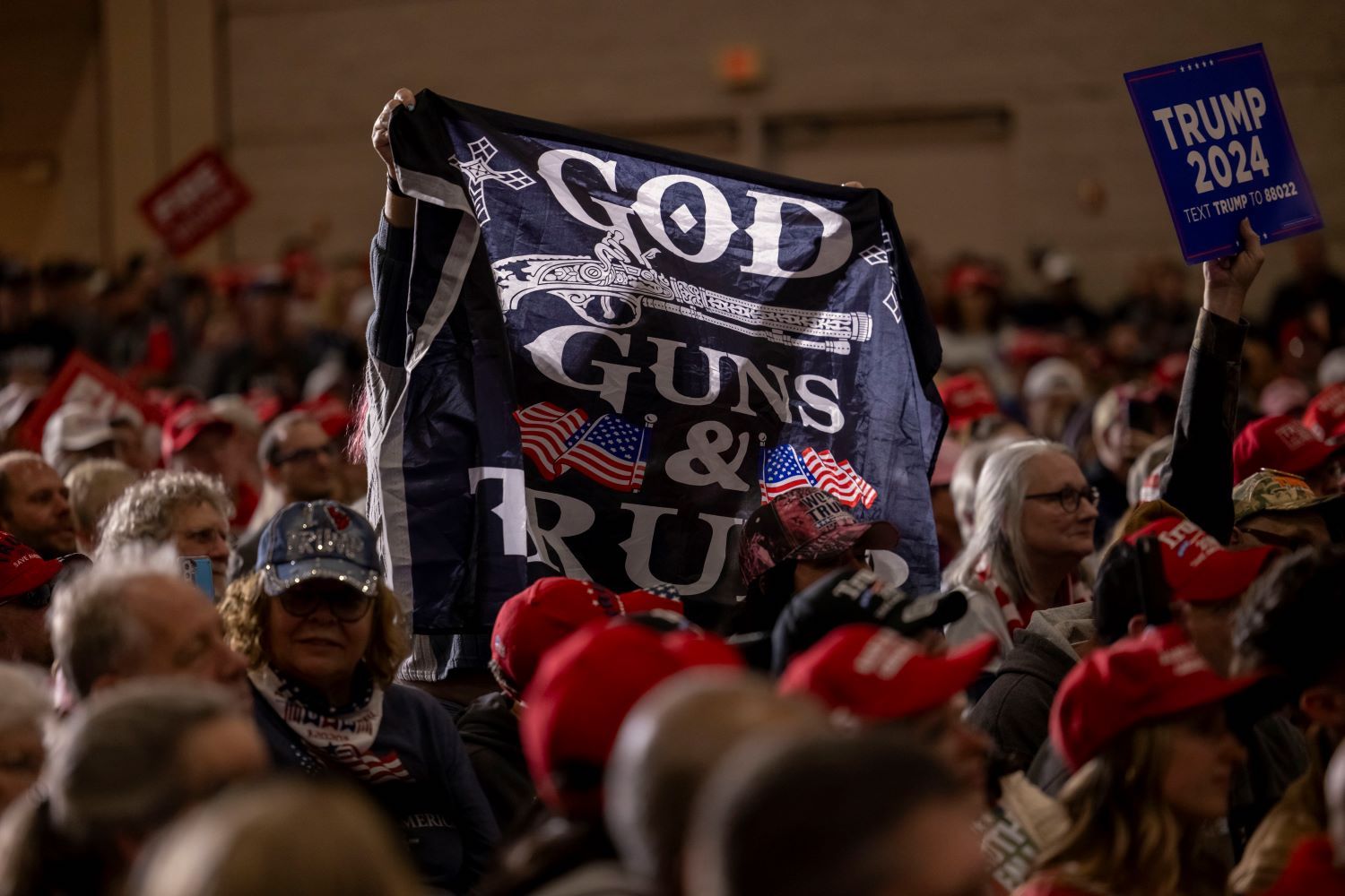 A crowd of people, many wearing red baseball caps. Someone holds up a fabric sign that reads 'God Guns & Trump.' Another sign reads 'Trump 2024.'