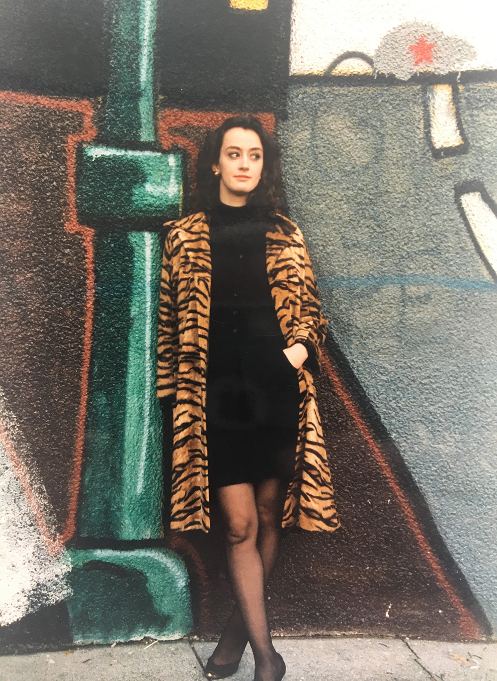 Dorothy Woodend stands against a graffiti wall in a short black dress, long tiger-print jacket, black tights and black shoes. She has long dark hair and is looking to the right.