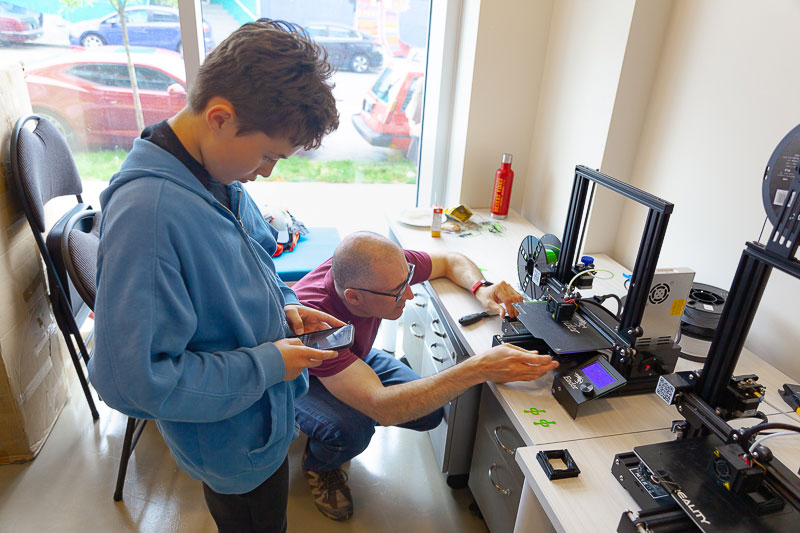 A youth looks at their phone as an adult crouches beside them and in front of a 3D printer.