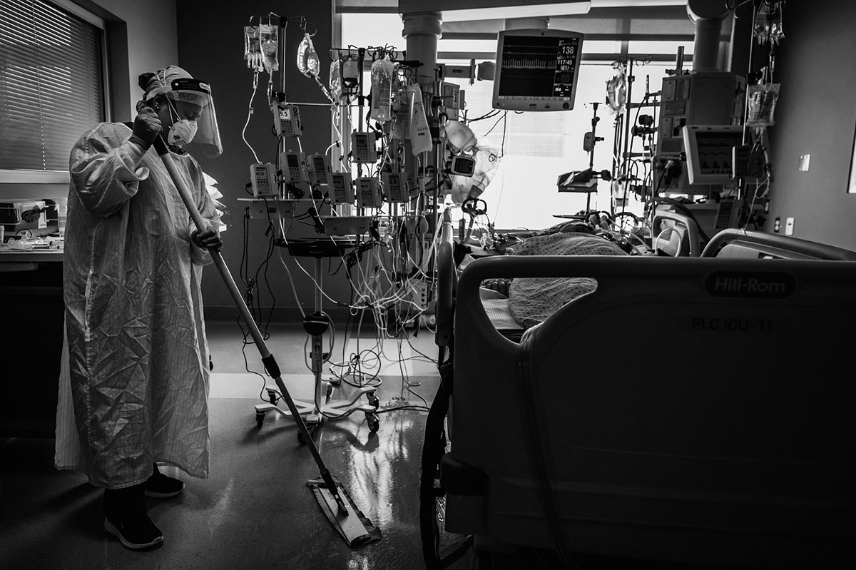 A woman in full protective equipment — her mask, her face shield, her gown — mops a hospital room. There is a patient in the bed to the right and over a dozen IV drips next to him.