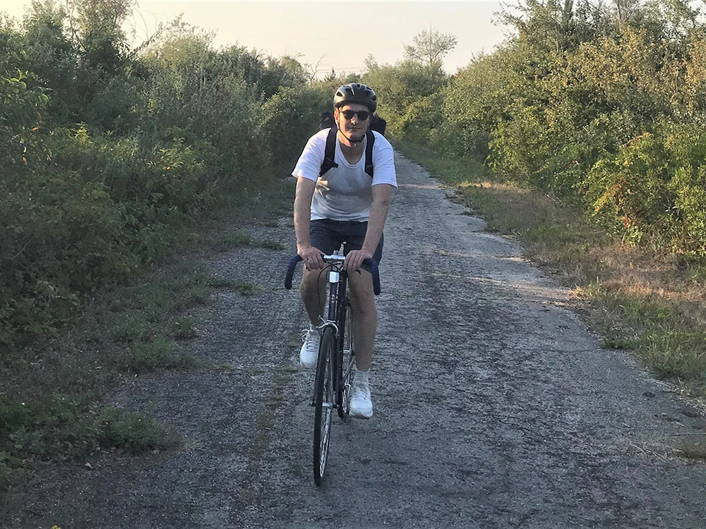 A man wearing a bike helmet and sunglasses rides a bike down an old paved path bordered by shrubs.