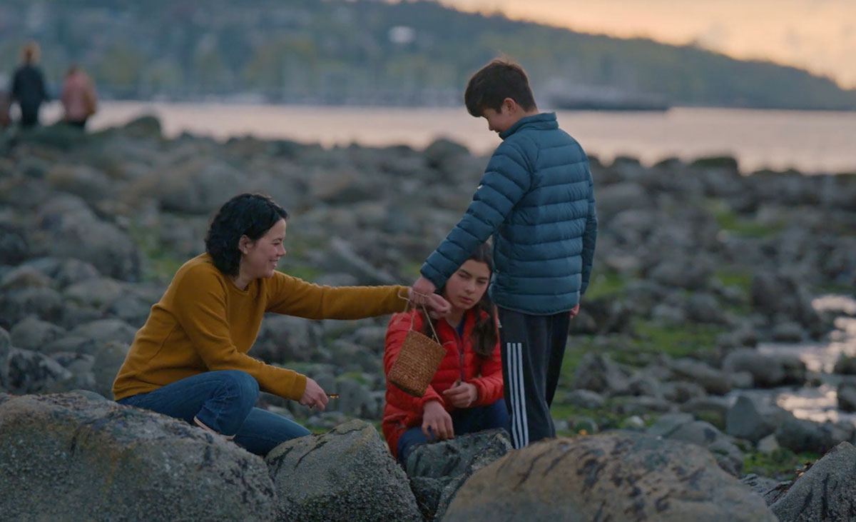 Three people are in the foreground of this image. On the left, a woman in a yellow sweater crouches amidst the large grey rocks on a beach and reaches towards a bucket that a young person in a puffy blue jacket is holding towards her. Between them, another young person in a red jacket is seated on the rocks and looking ahead, away from the camera. Behind the group are additional rocks across the beach, the ocean and a hill in the background.
