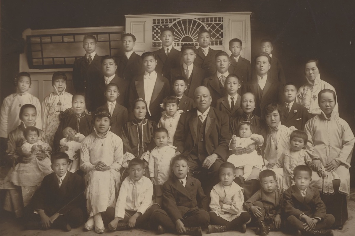 An old sepia photo of a large family of children and wives with a grandiose-looking man sitting at the centre.