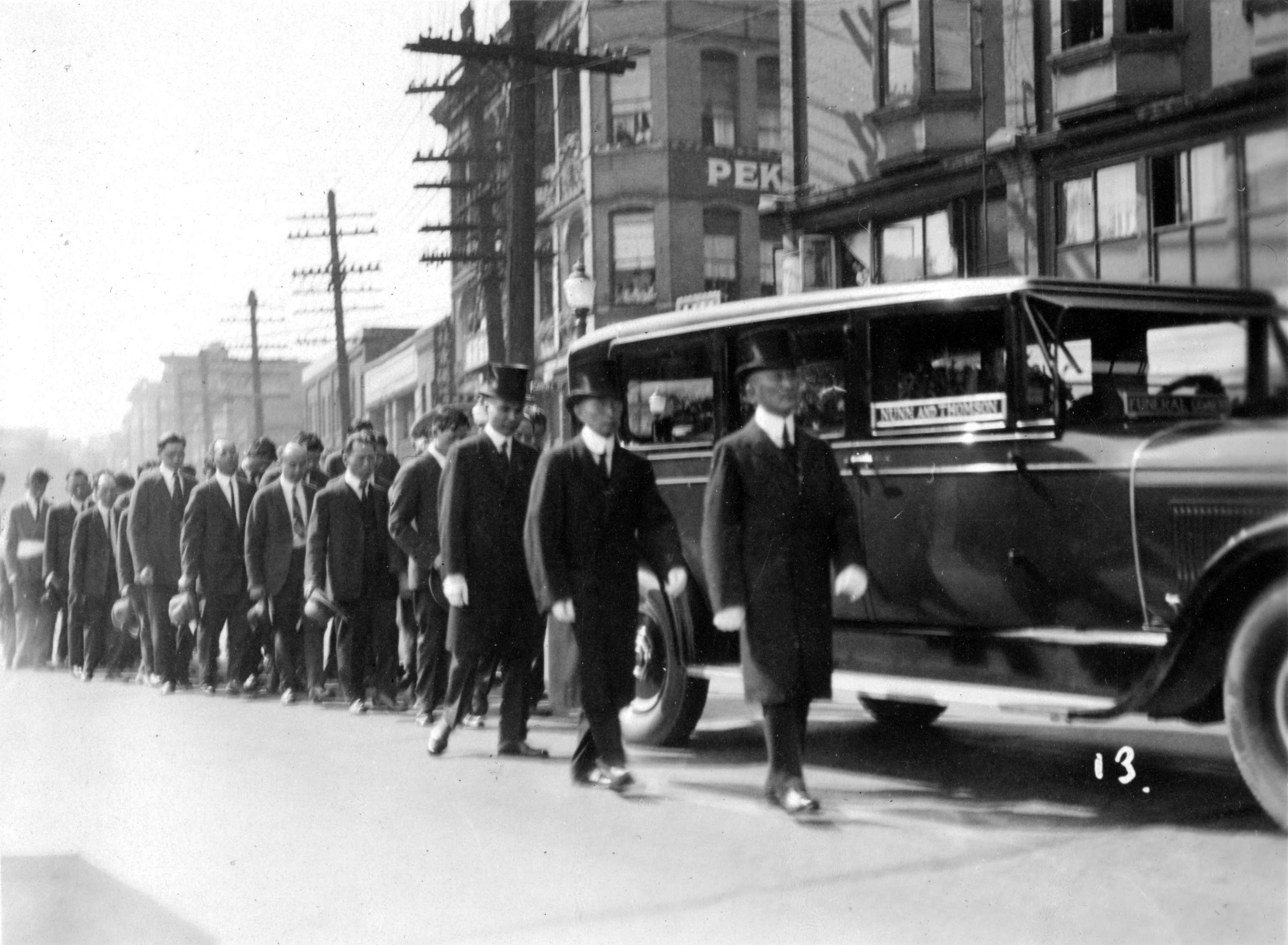 A black-and-white photo of a funeral profession with men in suits and top hats beside a hearse.