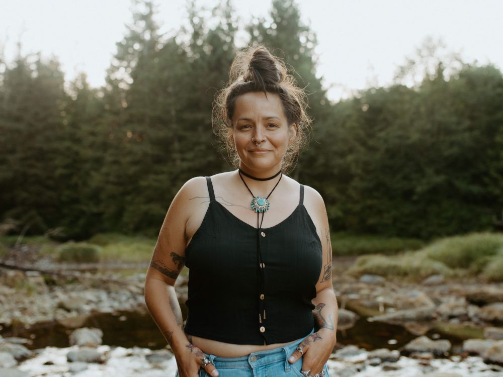 Jess Housty stands facing the camera, smiling. Their hair is tied in a topknot and they are wearing a black tank top with light blue jeans. Behind them is a stand of trees and a rocky river. 