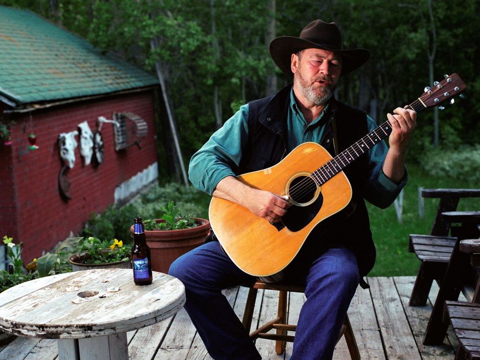 Sid Marty plays the guitar, wearing a dark brown cowboy hat, a black vest over a green long-sleeved shirt and jeans. He is outdoors on a wooden deck, with a beer bottle sitting on a rustic wooden table beside him. Green grass, trees and a red building with a green roof can be seen in the background.