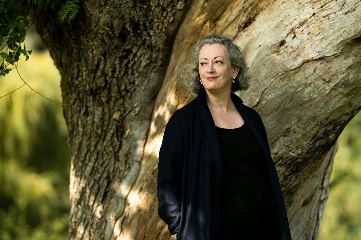 Dorothy Woodend stands against the wide trunk of a deciduous tree. She is wearing a long navy coat and has short, wavy grey hair. Greenery in soft focus fills the background.