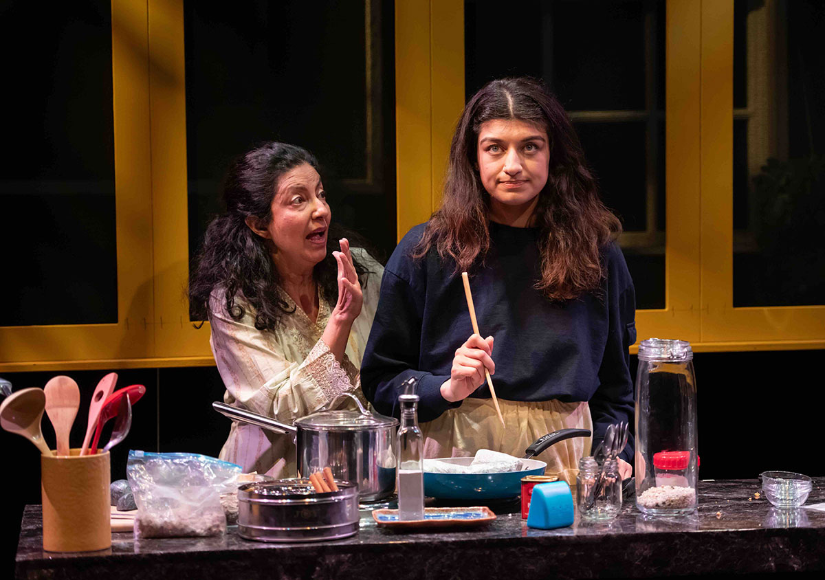 Two actors stand together behind a kitchen set onstage under diffused stage lights. In a light yellow dress, Nimet Kanji stands behind Talia Vandenbrink in a dark shirt. Vandenbrink stands over a pan holding a wooden utensil, rolling her eyes. 