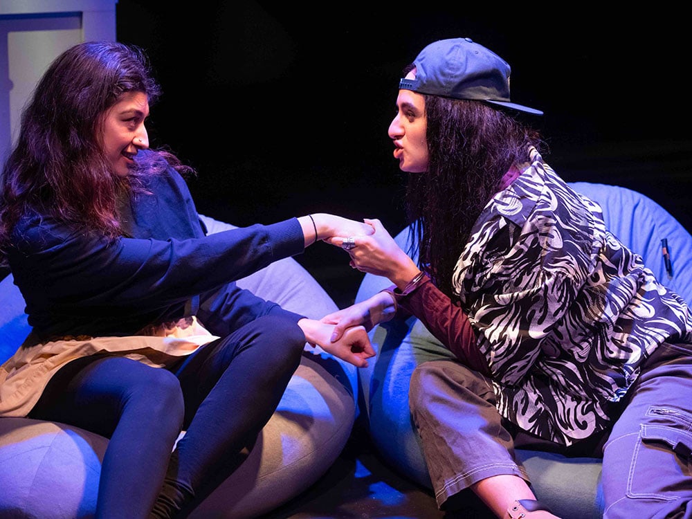 Two young actors interact with each other onstage under blue light while seated on bean bag chairs. The person on the right in a backwards baseball cap, a zebra-print shirt and cargo pants leans towards the person on the left, in dark denim and navy blue, taking their hand in a moment of shared conspiracy.