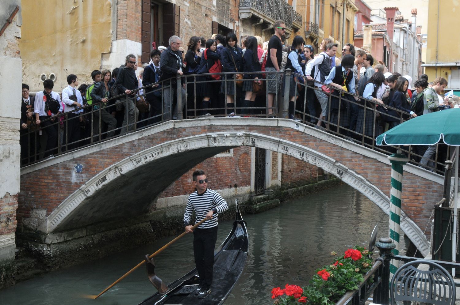 A throng of people on a small bridge over a canal in Venice. A gondolier in sunglasses and a white and black striped shirt poles a boat under the bridge.