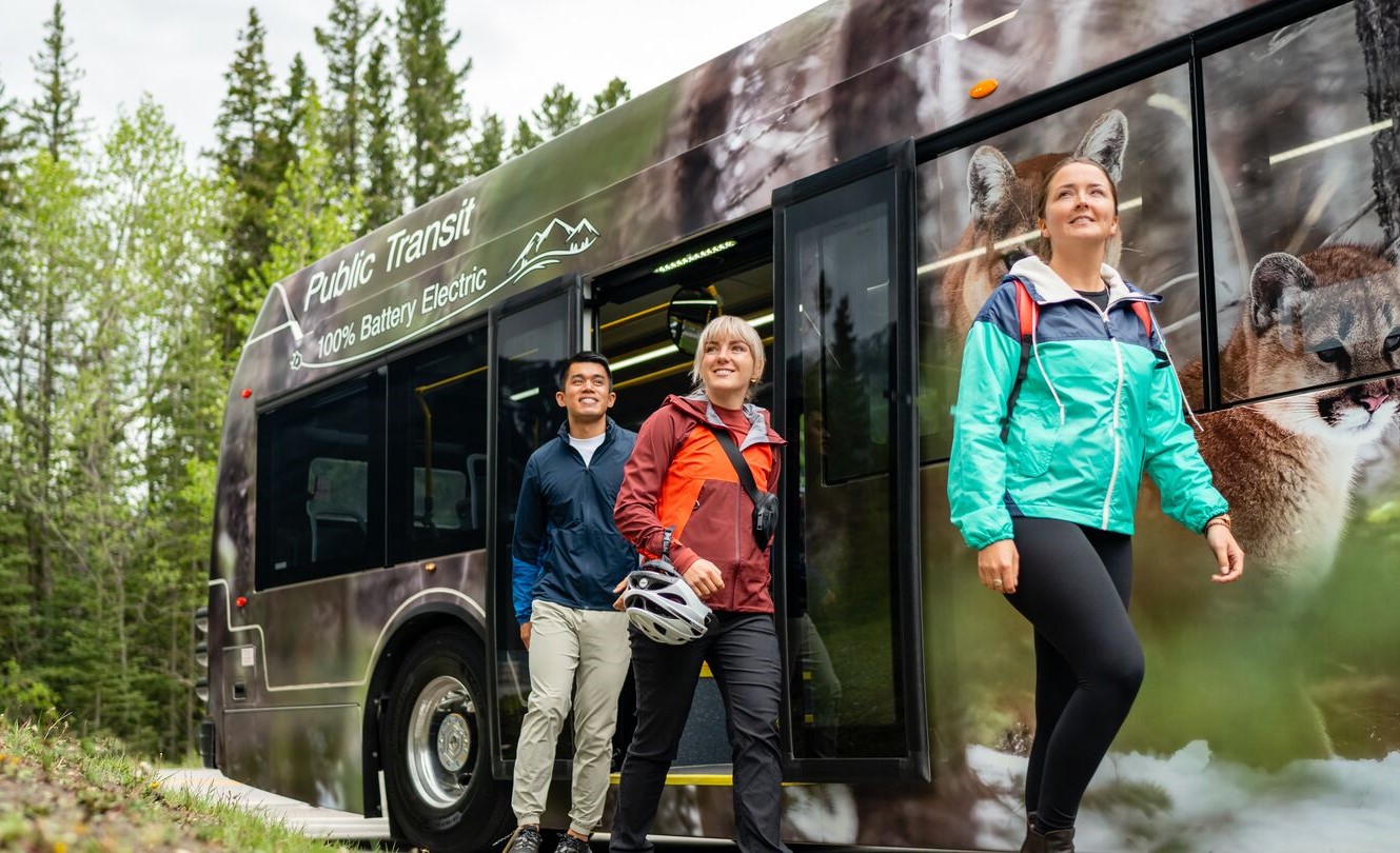 Three young adults in outdoorsy clothing disembark from a bus that says 'Public Transit 100% Battery Electric,' with trees in the background.