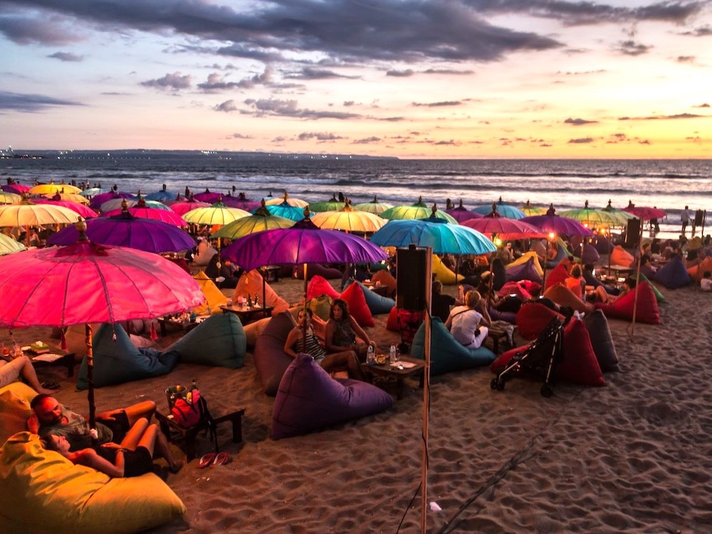 Colourful beach umbrellas cover people lounging in beanbag chairs on a beach at sunset.