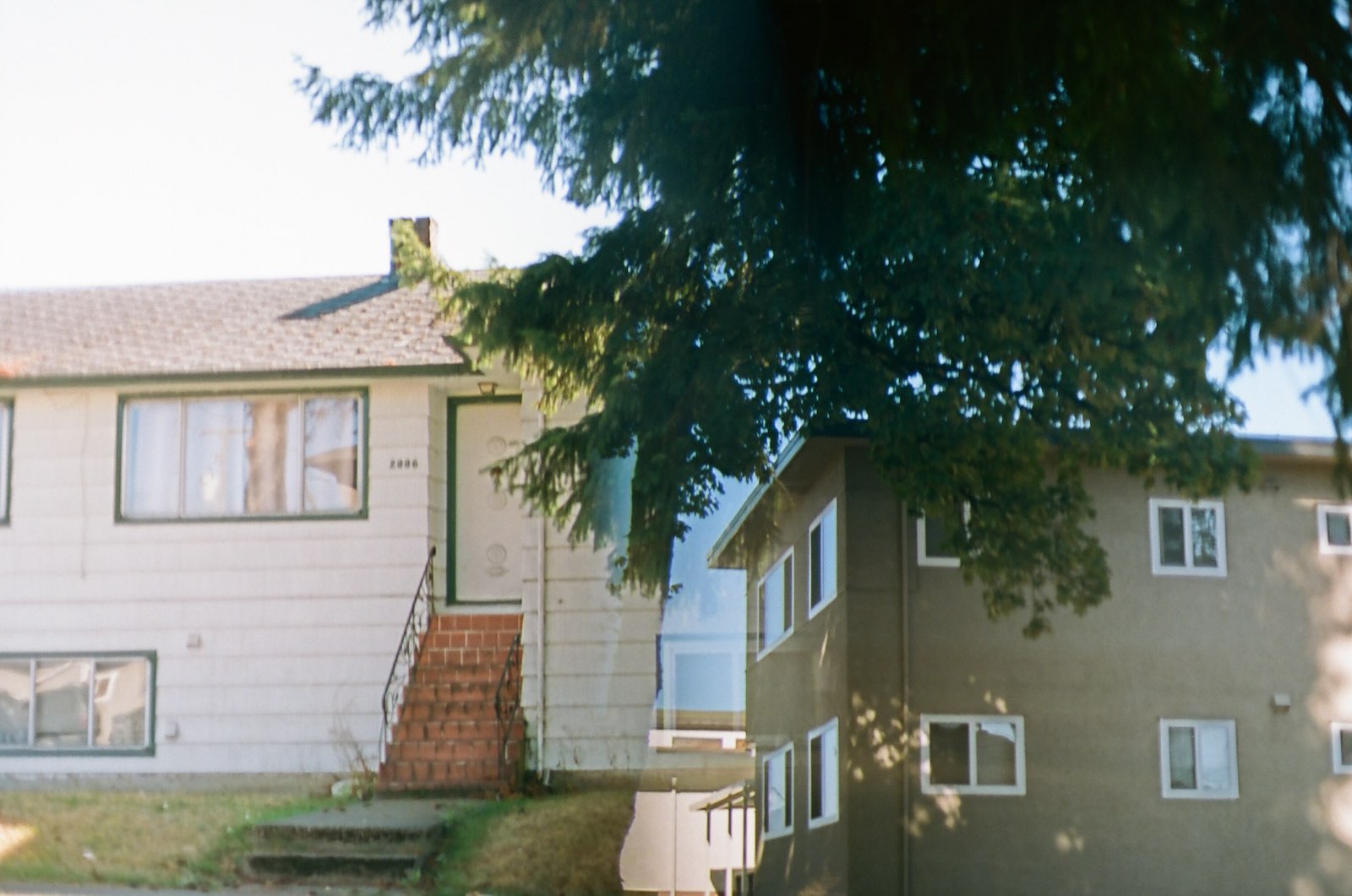 A double-exposure photograph depicts, on the left, a small white house, and, on the right, a grey three-storey walk-up apartment building, both on sunny days.