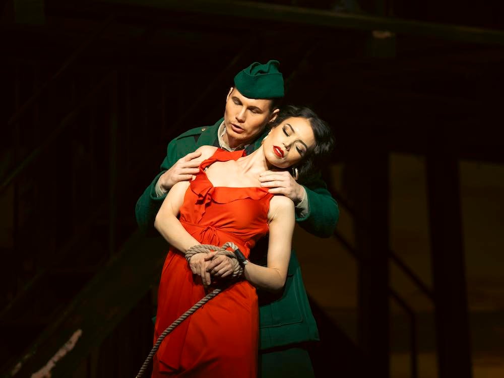 Two opera singers stand onstage against a darkened backdrop. Matthew White, left, has short hair, a green beret and light skin. He is clad in a green military costume. He holds the shoulders of Ginger Costa-Jackson, right, who leans against him with her eyes closed. She has light skin, dark wavy hair and dramatic makeup. She wears a red dress and her wrists are tied with ropes.