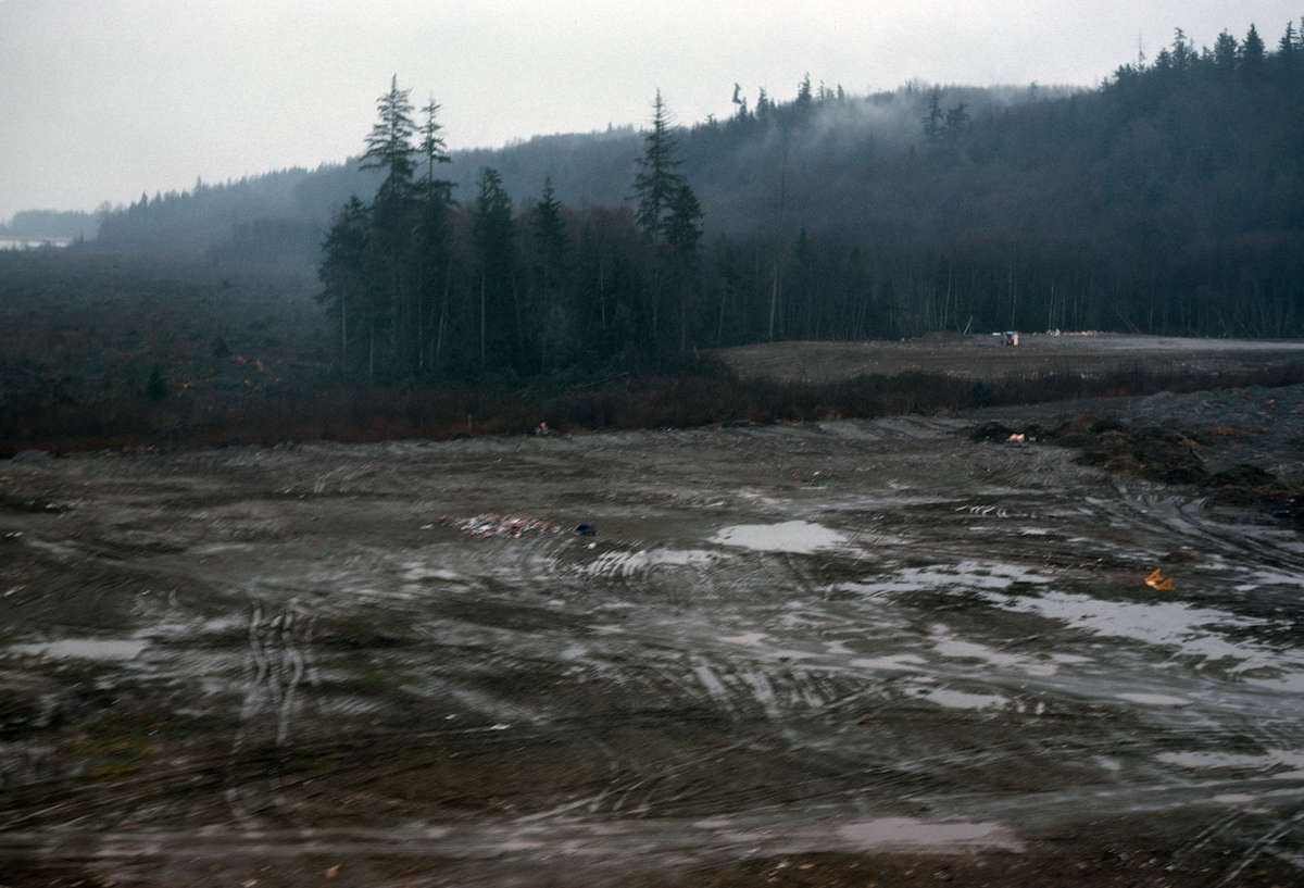 A photograph from 1976 showing a muddy site with pools of water and mounds of dirt. There is an orange barricade on its side. In the background are tall green trees and a thick fog.