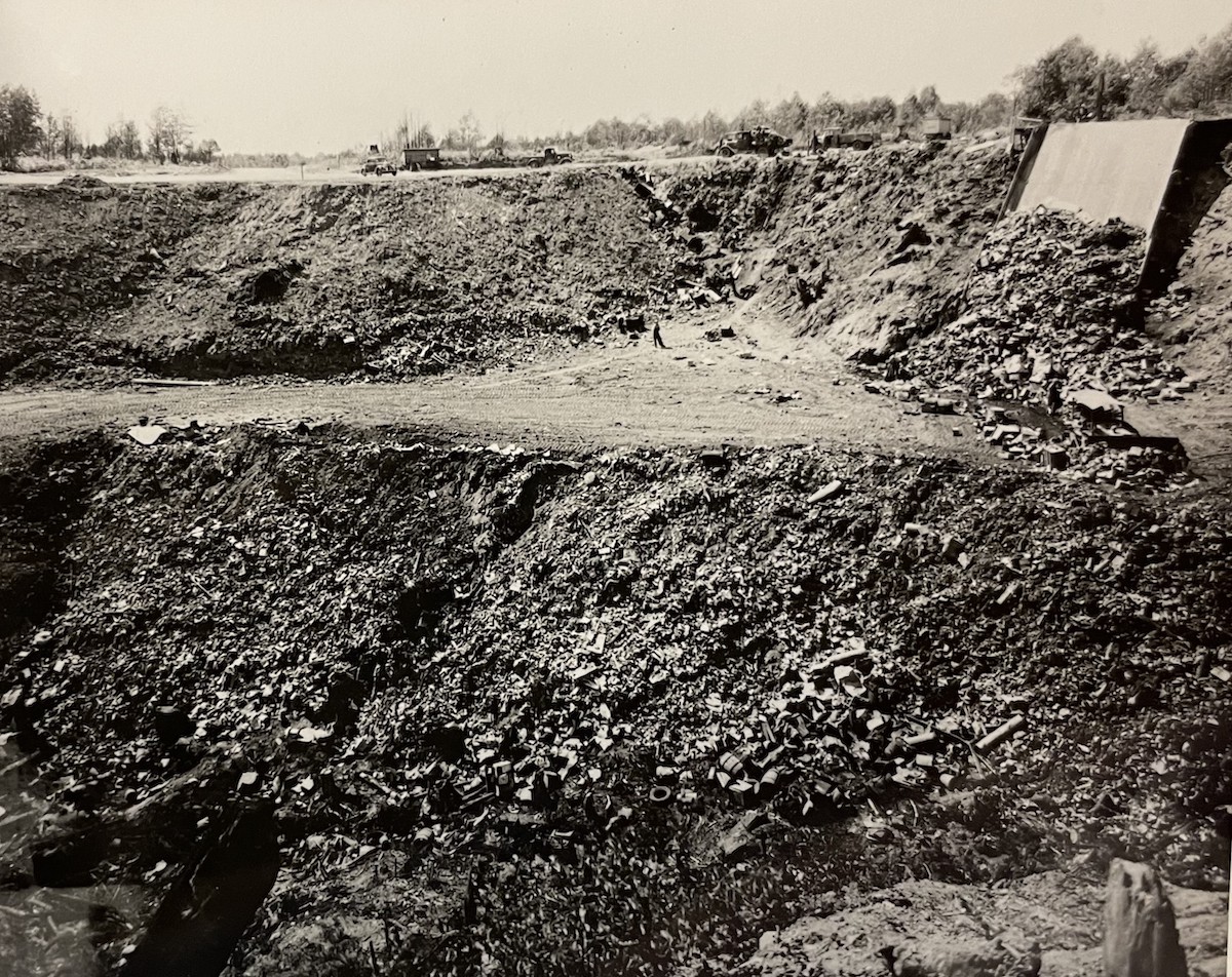 In a sepia-toned black and white photo, the figure of a person stands dwarfed by the enormous features of the Kerr Road Dump. There is a garbage chute to the right of the image, and a small structure on the hill above the figure. A big ditch is just across from the figure.