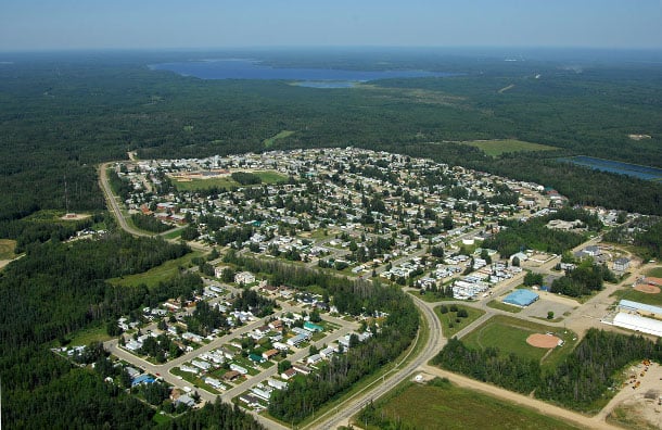 An aerial photo of Fox Creek. Houses are clustered along rows of streets. To the right, a river runs into a lake. The town, which seems relatively small, is surrounded by evergreen forest.