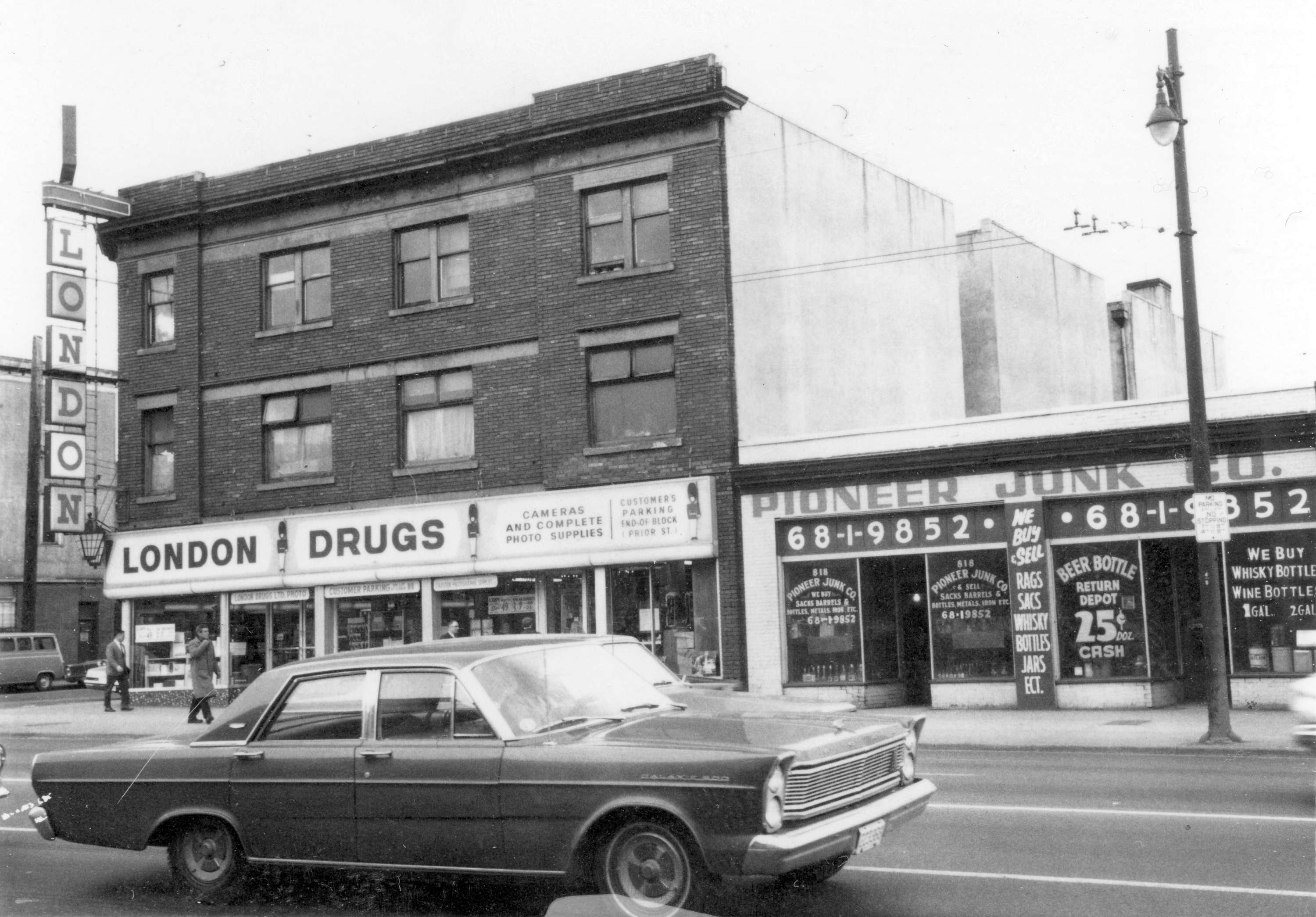 A black and white photo of Vancouver’s main street, taken in 1968. On the right is a one-story building home to the Pioneer Junk Co., with painted signs on the windows advertising bottle returns for cash. On the left is a three-story building, with a London Drugs store on the main floor. A sign reads, “Camera and complete photo supplies.”