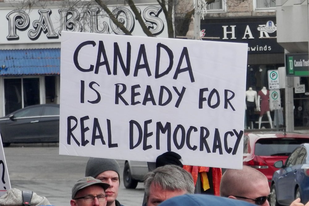 A protest sign held above a busy sidewalk says ‘Canada is ready for real democracy’ in handwritten black letters.