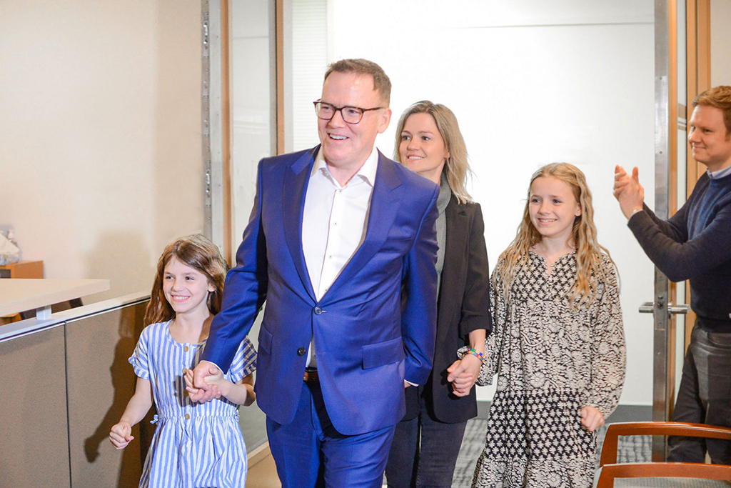 Kevin Falcon is wearing a blue suit and glasses. He is smiling and walking through a doorway into a room of supporters after winning the Vancouver Quilchena by-election in April 2022. He is holding hands with his youngest daughter, who is wearing a blue and white striped dress. Falcon’s spouse and oldest daughter walk behind him.