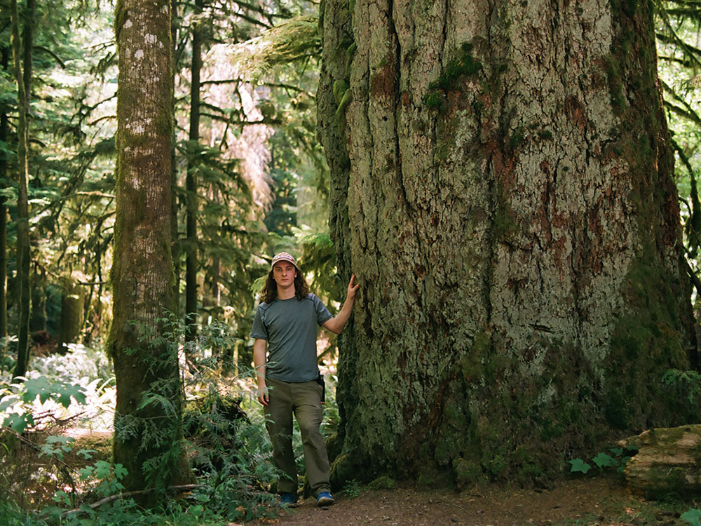A man with long hair stands next to a giant Douglas fir. He’s wearing a baseball cap.