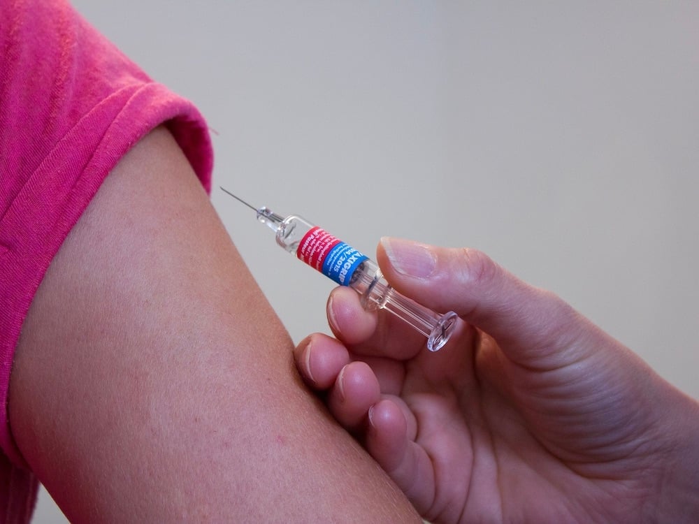 A syringe is poised to inject into the upper arm of a person wearing a pink T-shirt.
