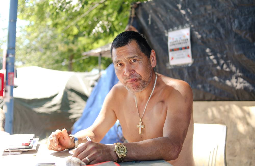 An Indigenous man with short hair, no shirt, a gold watch and a gold cross around his neck looks into the camera. He’s sitting at a table in the shade on a hot day.