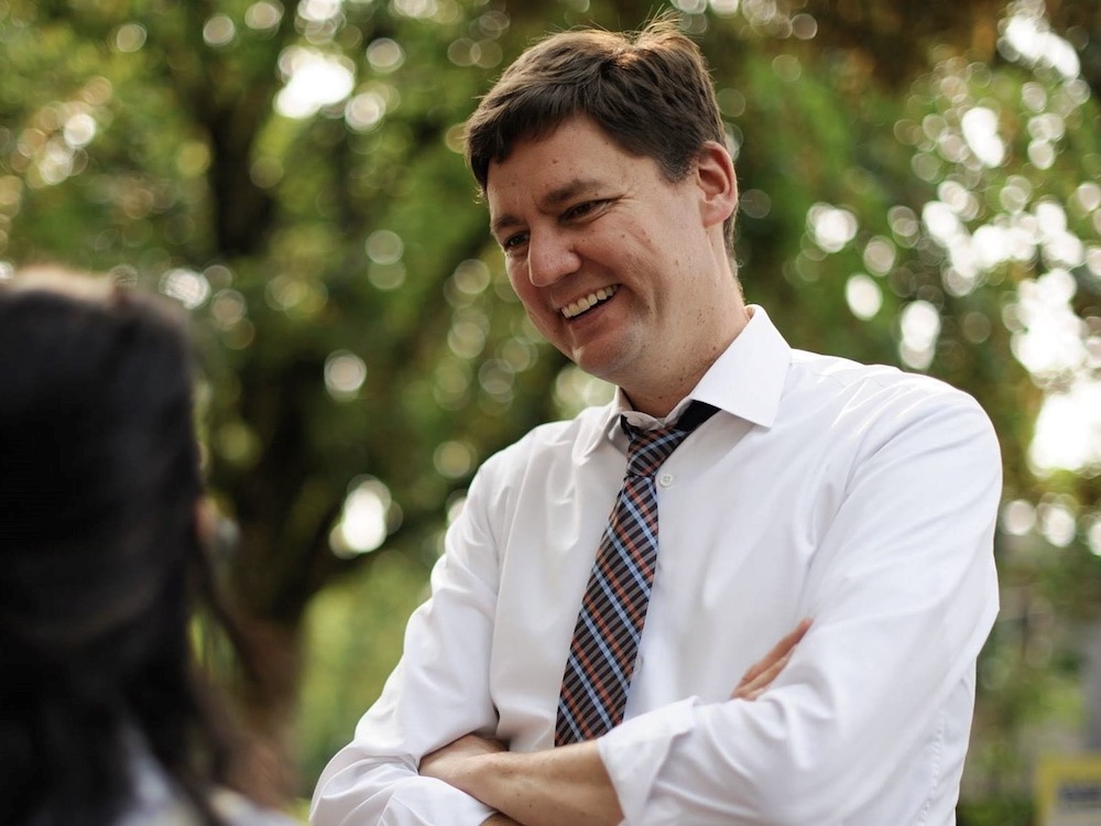 Brown-haired David Eby smiles outdoors in a white shirt and loosened plaid tie, speaking with a dark-haired female constituent against a backdrop of out of focus greenery.