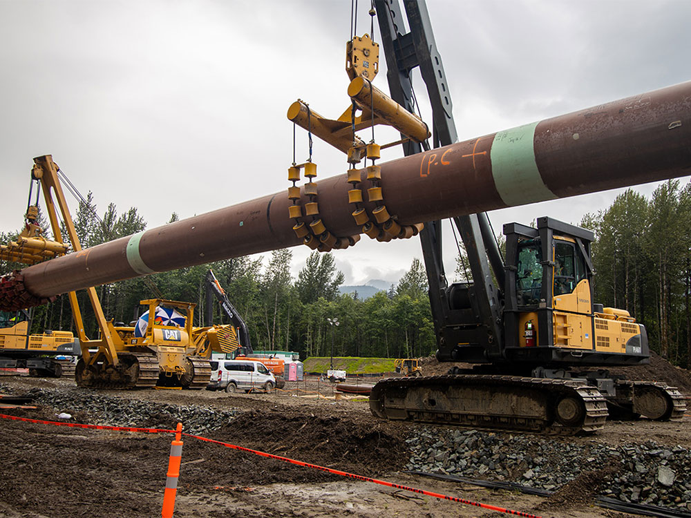 Two cranes attached to yellow bulldozers suspend a long stretch of pipeline above a bare patch of ground.