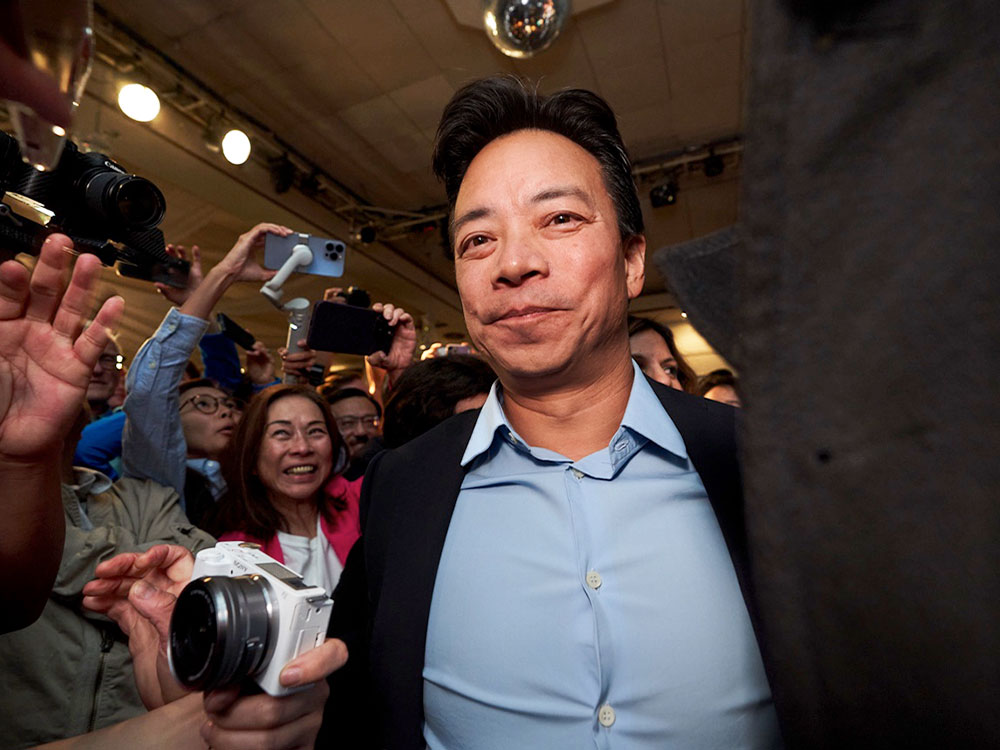 A closeup photo of Ken Sim, a middle-aged Chinese man. He is wearing a light blue button-up shirt under a black blazer. He is smiling with tears in his eyes. Crowded behind him are people smiling, cheering and taking pictures.
